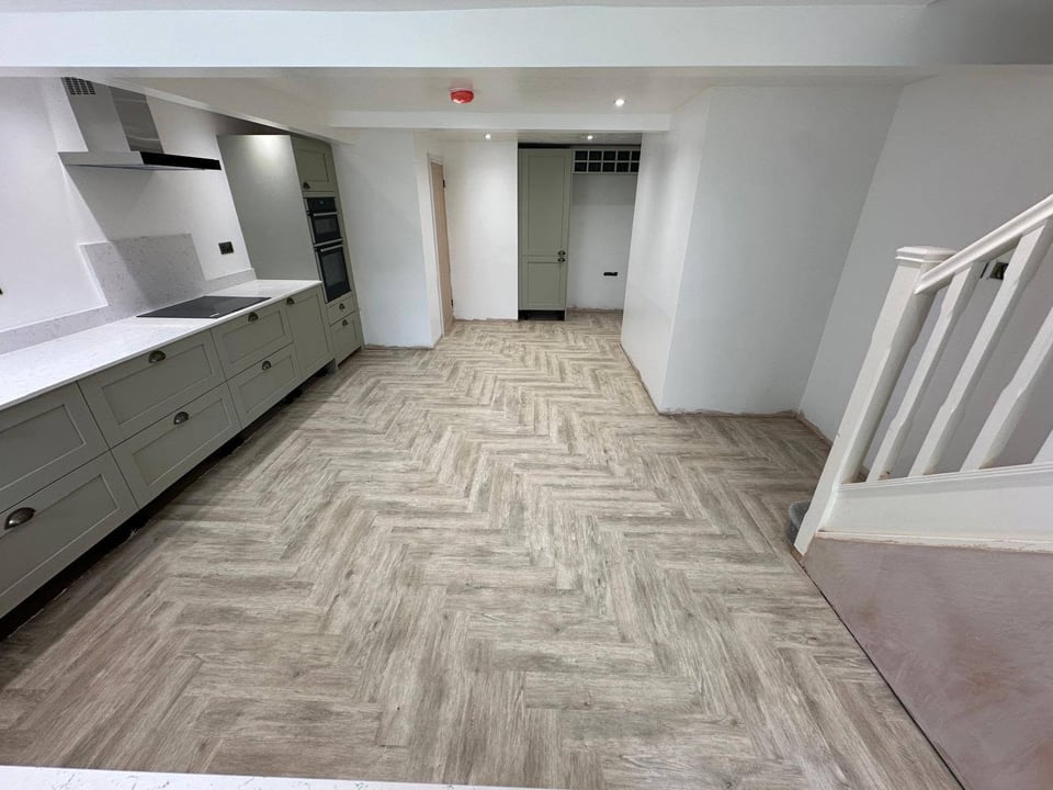 Long kitchen with grey cabinets, white worktop, herringbone floor and staircase