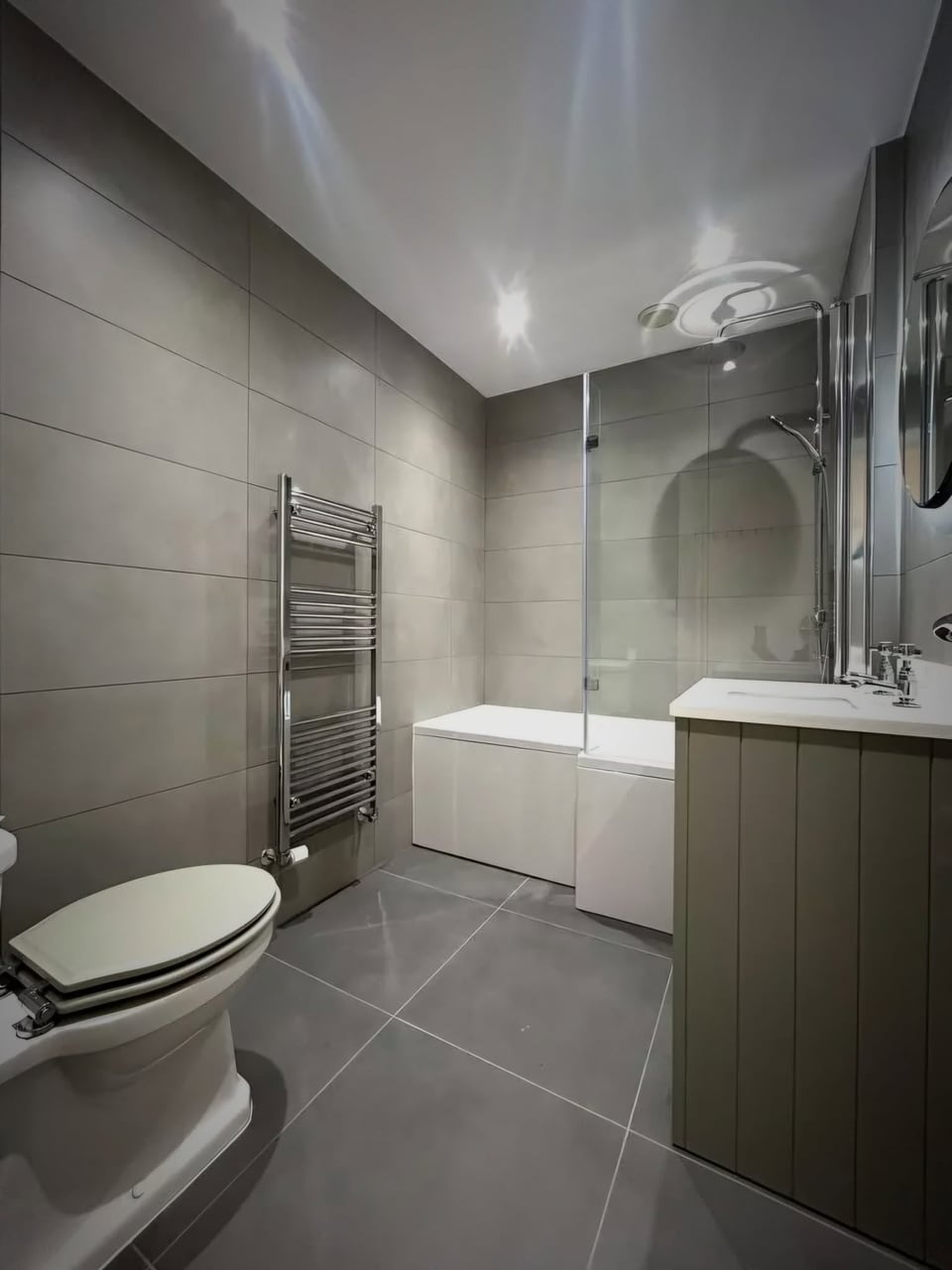 Grey-tiled bathroom with a shower over the bath, chrome towel rail and dark vanity unit